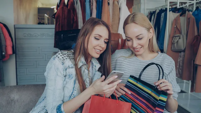 Two women enjoying a shopping spree and sharing a moment on their smartphone in a clothing store. Implementing a retail media tool from release boundary to operable handover